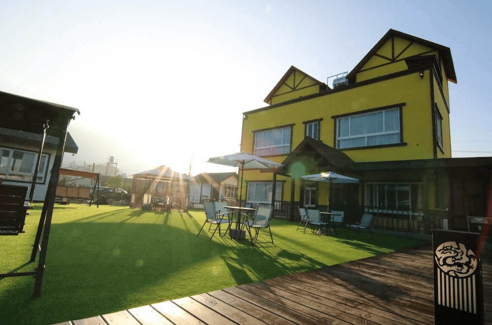 【埔里住宿】埔里日記的民宿外觀以亮黃色牆面為主，搭配綠意盎然的庭院草坪，庭院中設有戶外座椅與遮陽傘，陽光灑在草地上，整體呈現歡樂與放鬆的渡假感。