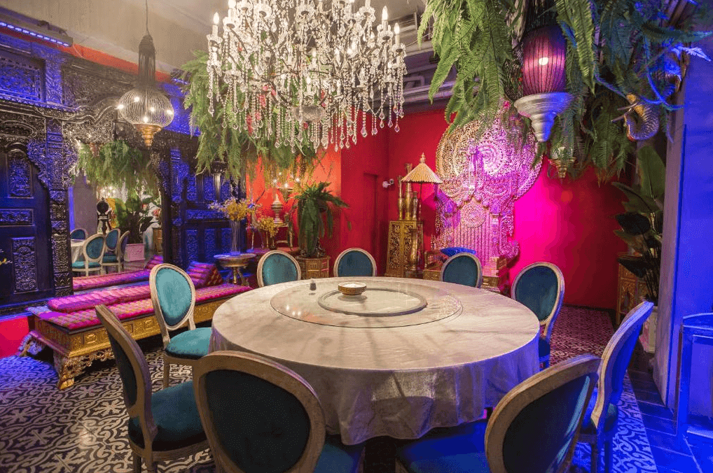 【嘉義寵物友善住宿】安娜國王酒店的東方宮廷風格的餐廳，擺放圓形大理石餐桌，周圍配有藍綠色絲絨椅子，天花板吊掛華麗水晶吊燈，搭配植物裝飾，背景牆上有精緻雕刻裝飾。