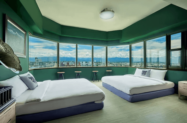 【嘉義寵物友善住宿】安蘭居旅店的全景窗戶現代化客房，牆面採用深綠色設計，內有兩張大床，窗外可欣賞城市景觀與遠山，房內佈置簡約而舒適。