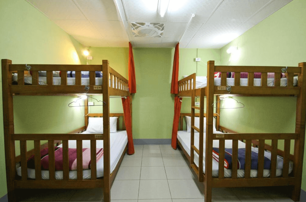 【埔里住宿】島中央旅人聚場的背包客多人房，設有兩排木質雙層床，每個床位配有白色床單和枕頭，房間牆面為淡綠色，並設有橘色窗簾增加隱私性，燈光柔和，營造溫馨氛圍。