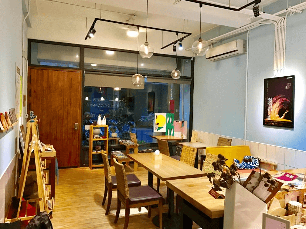 【埔里住宿】島中央旅人聚場的公共空間，設有木質桌椅，燈光溫暖柔和，牆面掛著多幅藝術裝飾，提供旅人交流與放鬆的環境，整體氛圍舒適且充滿設計感。