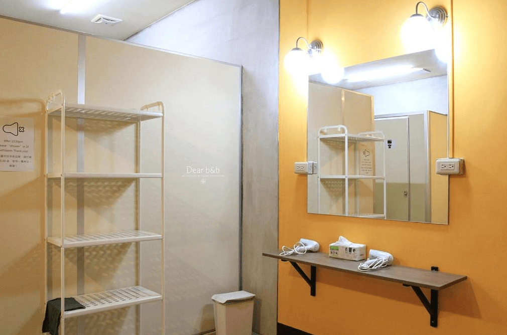 【埔里住宿】島中央旅人聚場的公共衛浴空間，牆面塗為明亮的黃色，配有大型鏡子和簡約的白色架子，提供吹風機與紙巾，環境整潔且實用，燈光充足。