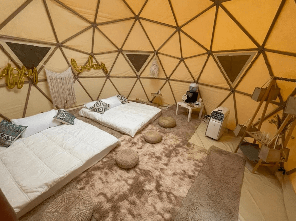 【司馬庫斯住宿】悠野_s露營區的圓頂帳篷內部，鋪設了兩張日式地墊床鋪，床上擺放著幾何圖案靠枕，地面鋪有柔軟地毯和藤編坐墊，牆上裝飾著流蘇掛飾和金色氣球，右側設有飲水機與冷氣機等設施。