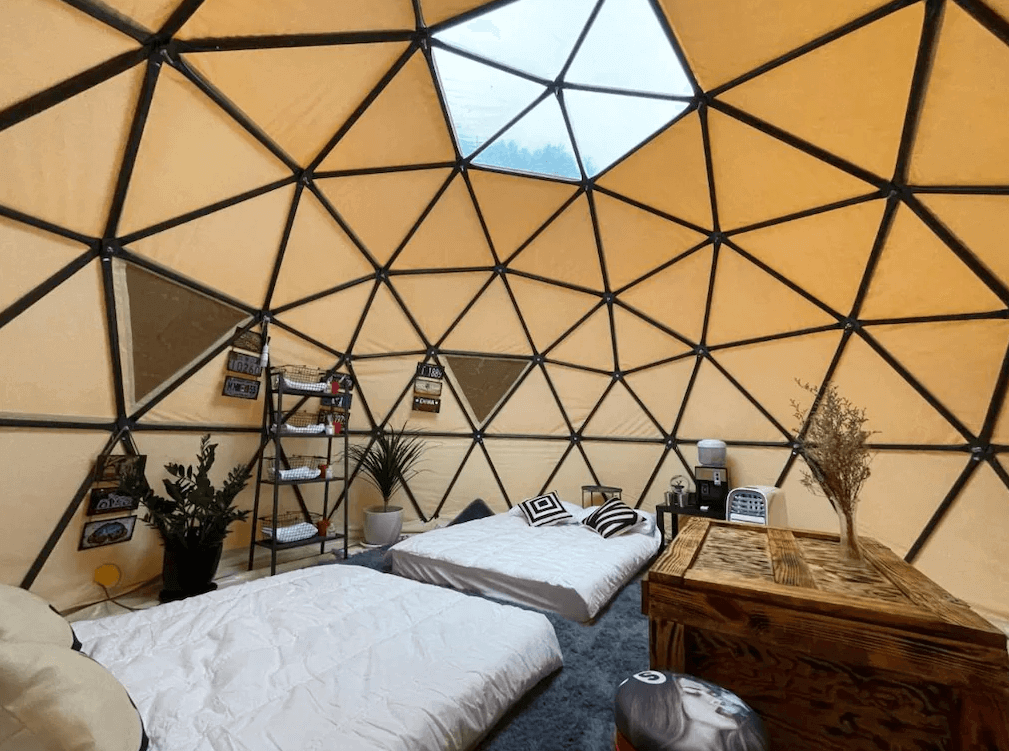 【司馬庫斯住宿】悠野_s露營區的圓頂帳篷內部空間，擺設了兩張床鋪，搭配簡約黑白靠枕，窗外透入自然光線。房內設有木製桌子、小型植物和簡約置物架，營造出舒適與自然結合的氛圍。