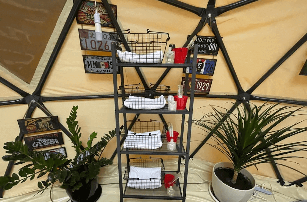 【司馬庫斯住宿】悠野_s露營區的圓頂帳篷內的一個置物架，上面整齊擺放著毛巾、盥洗用品和紅色杯子，周圍裝飾有復古車牌與綠植，整體呈現出整潔又有設計感的風格。