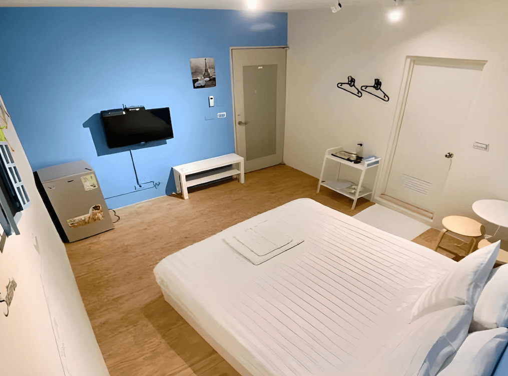 【新竹火車站住宿】懶骨頭 Lazy Bone的房間內裝潢簡約，以藍色牆面搭配白色床品，整體風格清新明亮。設有一台壁掛式電視、小型冰箱以及白色木質家具，為旅客提供基本的實用設施。地板採用木質紋理，增添溫馨感。