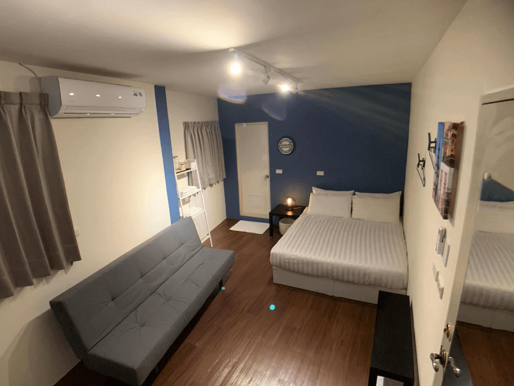 【新竹火車站住宿】懶骨頭 Lazy Bone的房間內設有一張雙人床和一張灰色沙發，提供靈活的住宿選擇。牆面以藍白色調為主，營造簡潔的視覺效果。室內配備空調、簡易衣架和小型家具，滿足旅客基本需求。