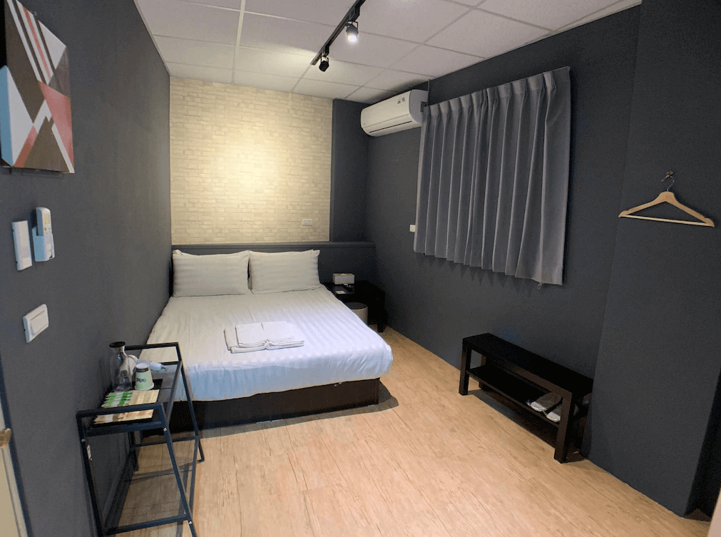 【新竹火車站住宿】懶骨頭 Lazy Bone的房間內牆面為深灰色，搭配一面淺色磚牆，營造現代簡約的風格。雙人床配有白色床品，設有衣架和簡易家具，空間佈局精緻實用，並提供舒適的住宿體驗。