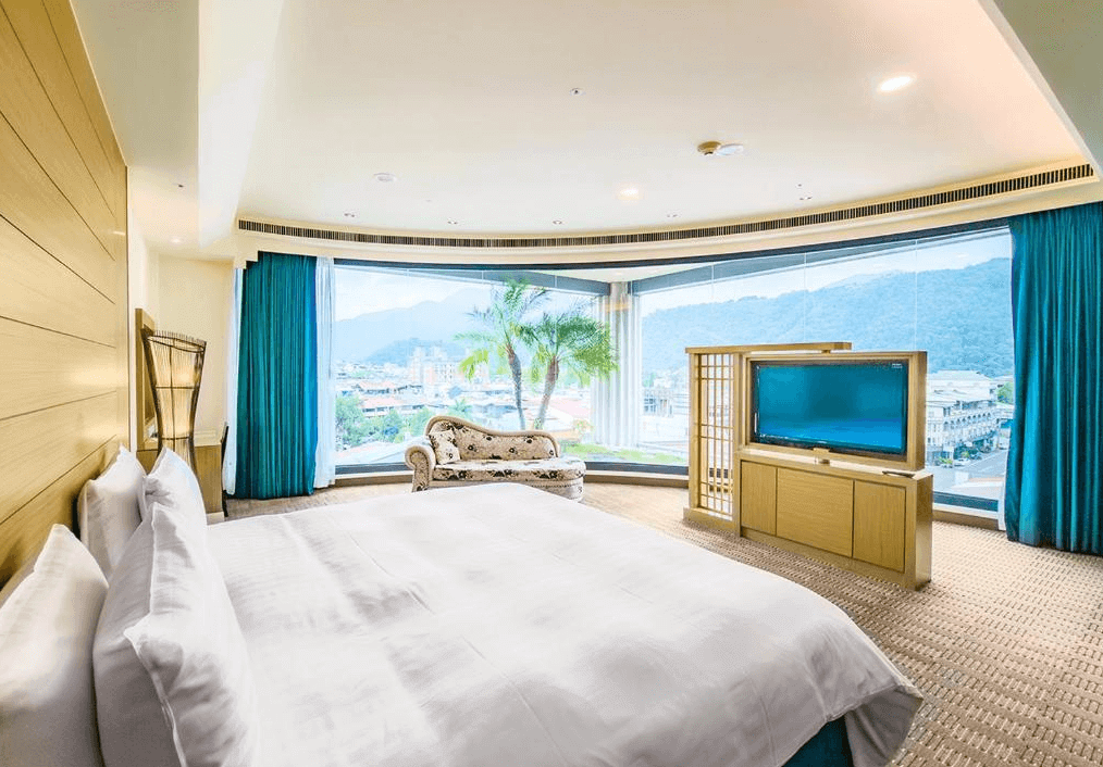 【埔里住宿】承萬尊爵渡假酒店豪華的客房，內部設有一張大床，床頭牆面採用木質裝飾。房間擁有寬敞的落地窗，可以欣賞窗外青山與城鎮的景緻。窗邊還擺放著一張復古風格的沙發椅，整體感覺溫馨又舒適。