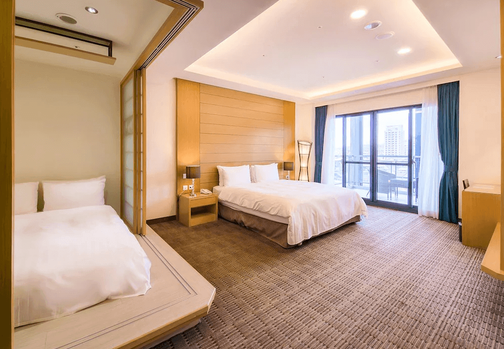 【埔里住宿】承萬尊爵渡假酒店的家庭友善的客房，內部擁有兩張床，其中一張設置在滑動門隔開的小空間內，營造隱私感。主臥區域設有陽台，透過玻璃門可以俯瞰外部景色。房間以木質和暖色調設計，氛圍溫暖舒適。