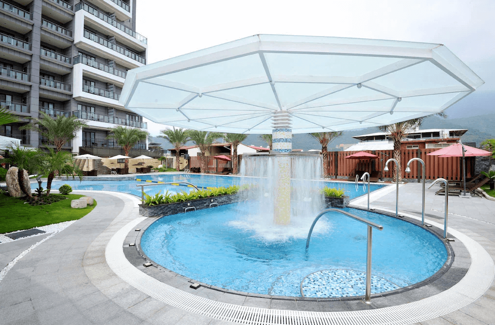 【埔里住宿】承萬尊爵渡假酒店的戶外泳池區，設有水柱與遮陽傘，泳池旁環繞著綠植與棕櫚樹，營造熱帶渡假氛圍。泳池邊還有休憩的躺椅與遮陽棚，適合享受放鬆時光，背景可見飯店建築與青山景色。