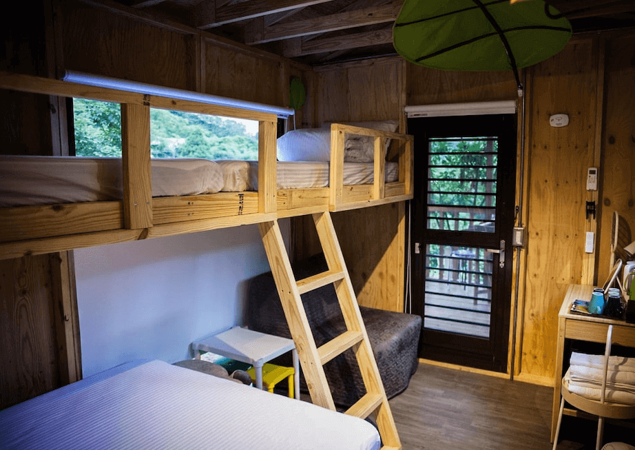 【司馬庫斯住宿】拾階莊園（內灣露營車）的雙人房，床上鋪有潔白的寢具，房間內設有平板電視、簡約書桌與椅子，推拉玻璃門通往陽台，光線明亮，窗簾為灰色，地板採用木紋設計。