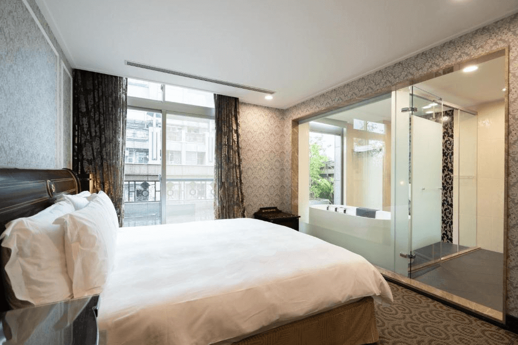 【新竹火車站住宿】新舍商旅新竹館的房間內設有一張大型雙人床，背景牆以灰色浮雕壁紙裝飾，呈現典雅風格。房間內浴室採用透明玻璃設計，可看到大型浴缸，搭配窗外的綠色景觀，整體感覺開放且舒適。