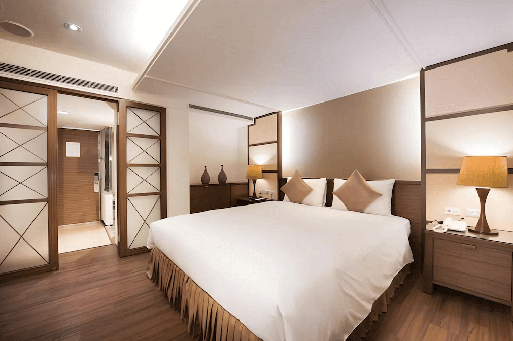 【新竹火車站住宿】新苑庭園大飯店的房間內設有一張大雙人床，床尾搭配兩盞暖色調床頭燈，營造溫馨氛圍。木質地板與家具設計簡約大方，房間背景牆採用米色搭配木質元素，突顯現代優雅感。