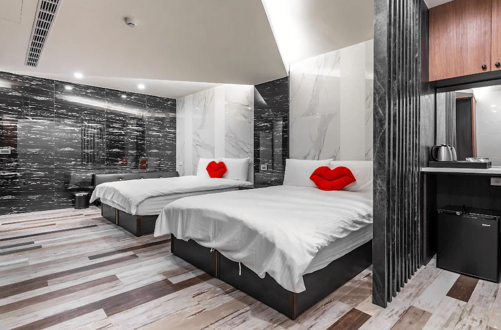【日月潭住宿】日月潭兒茶宿的房間以大理石紋牆面搭配亮面材質設計，展現出時尚摩登的風格。雙人床設計搭配紅唇抱枕點綴，增添趣味性與溫馨感。地板採用多色木紋拼貼，營造溫暖氛圍，並設有簡易小吧檯及冰箱，提供便利的住宿體驗。