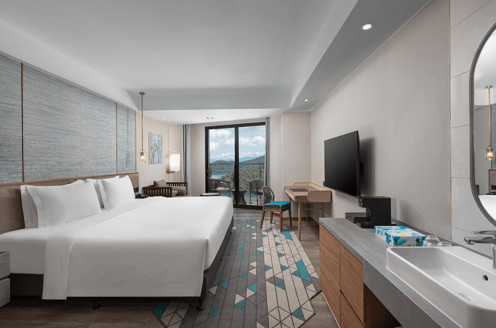 【日月潭住宿】日月潭力麗溫德姆溫泉酒店的房間內配有寬敞的雙人床，佈置以淡雅色系為主，搭配室內懸吊燈飾，展現現代簡約風格。落地窗通往陽台，可享受湖景與戶外空氣。