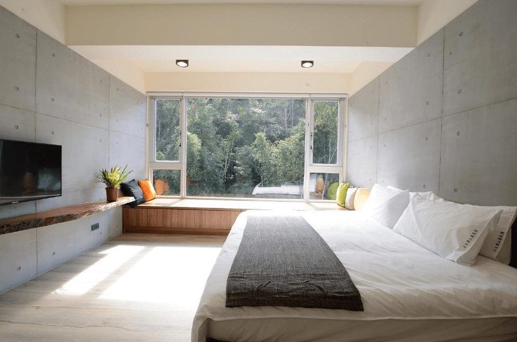 【日月潭住宿】日月潭山慕藝旅的房間採用現代極簡風格設計，整體以清水模牆面搭配溫暖木質元素，窗邊設有舒適的窗台座椅，視野開闊，可欣賞窗外綠意盎然的自然景色。大床鋪陳簡約，搭配柔軟的枕頭與寢具，營造出放鬆的住宿氛圍。