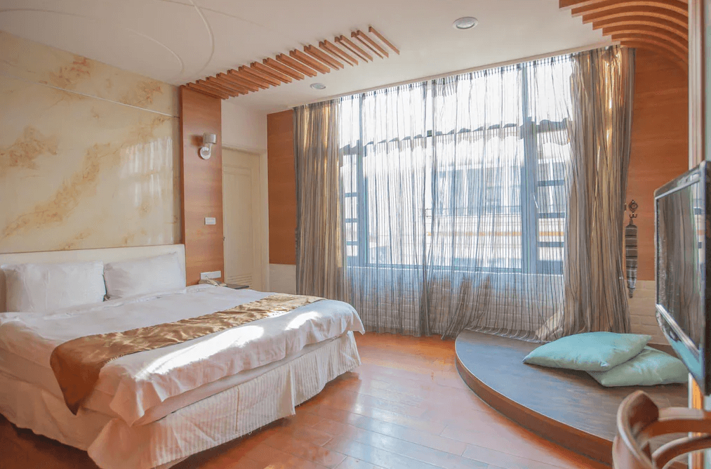 【日月潭住宿】日月潭水岸休閒飯店溫暖木質調的房間，床旁有落地窗，陽光灑入，窗簾優雅地垂下，旁邊設有小圓桌與墊子，營造舒適的居家氛圍。