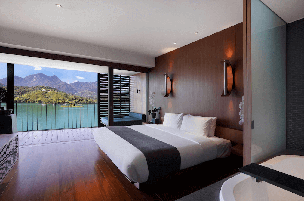 【日月潭住宿】日月潭涵碧樓酒店的房間內擁有一張大床，背景以木質牆面點綴，旁邊是全景落地窗，外可見湖光山色，環境寧靜優雅。