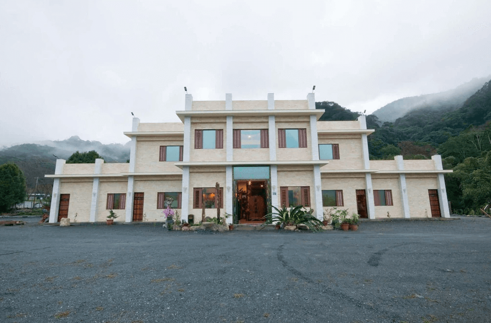 座落於群山環繞中的春陽一品居，有兩層樓建築，外觀簡約大氣，白色與米色調的外牆搭配對稱窗戶設計，散發典雅氛圍。前方為寬敞的停車空間，周圍被綠意盎然的山景環抱，展現寧靜悠閒的自然環境。