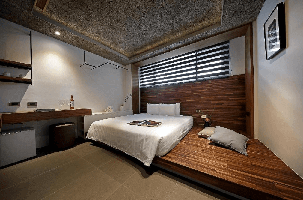 【日月潭住宿】桂月村的房間內使用深色木質元素與白色牆面搭配，展現簡潔且溫暖的氛圍。床旁設有貼地木質座位區，增添實用性與設計感，窗戶搭配調光窗簾，提供明亮與隱私兼具的空間。