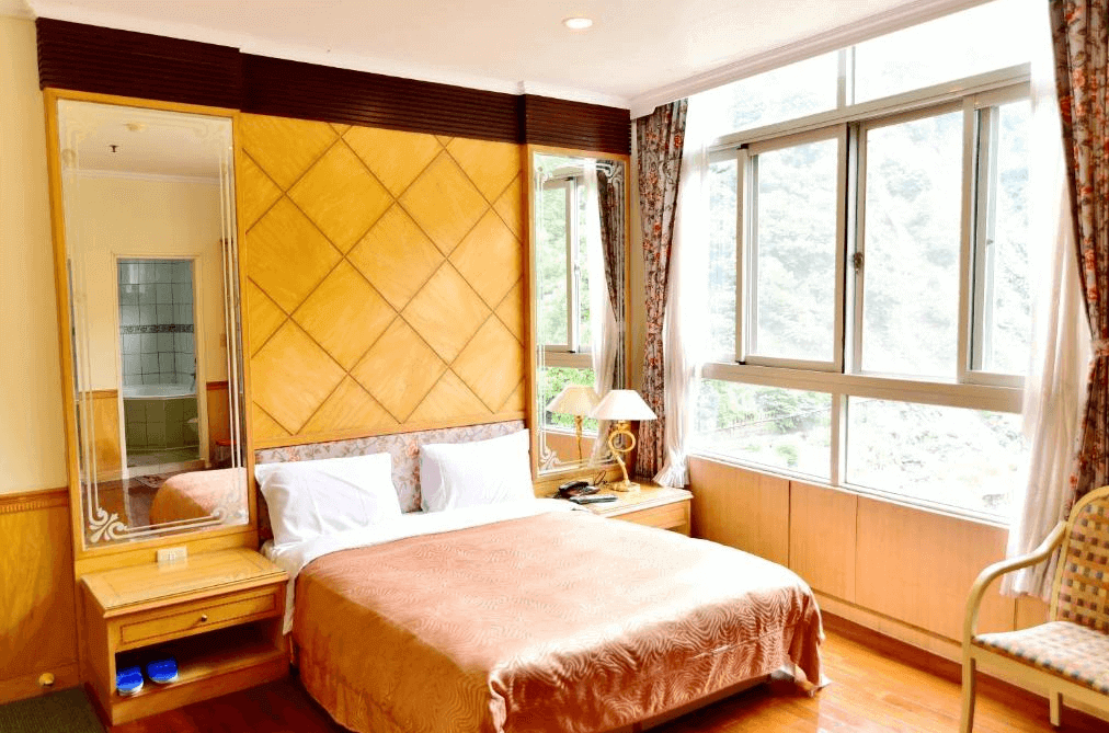 【廬山溫泉】正揚大飯店採光良好的雙人房，擁有大片落地窗，可俯瞰山林景色。房內裝潢以木質與暖色調為主，搭配經典格紋牆面設計與花紋窗簾，營造出典雅舒適的氛圍。房內備有床頭燈、梳妝鏡與小茶几，提供便利的住宿體驗。
