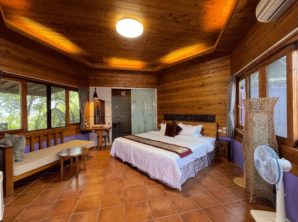 【埔里住宿】水月山莊的木屋風格客房，室內以原木裝潢為主，搭配白色床鋪和紫色布藝沙發，地板採用溫暖的紅磚色調，窗外可見自然景觀，營造放鬆的渡假氛圍。