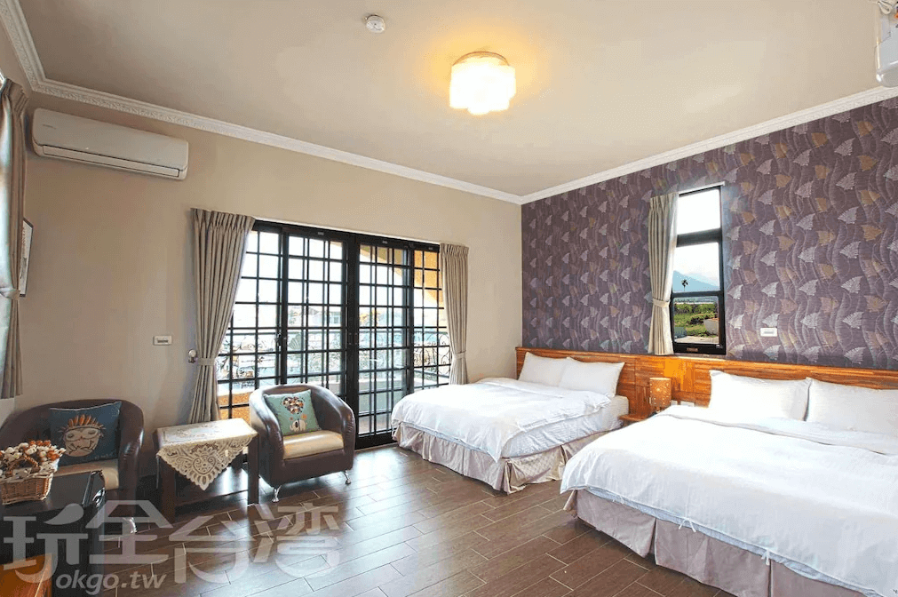 【埔里住宿】水部子山莊民宿的家庭房，內有兩張雙人床，牆面採用紫色幾何圖案壁紙裝飾，增添現代感。房間設有落地窗與陽台，陽光充足，角落擺放兩張皮椅與小桌，適合休憩，整體氛圍溫暖舒適。