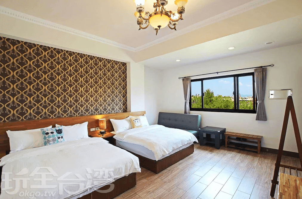 【埔里住宿】水部子山莊民宿的雙床房，牆面設計為金色幾何圖案壁紙，搭配暖色調燈光。窗外景色綠意盎然，室內家具包含木質床架與灰色沙發，空間明亮且舒適。
