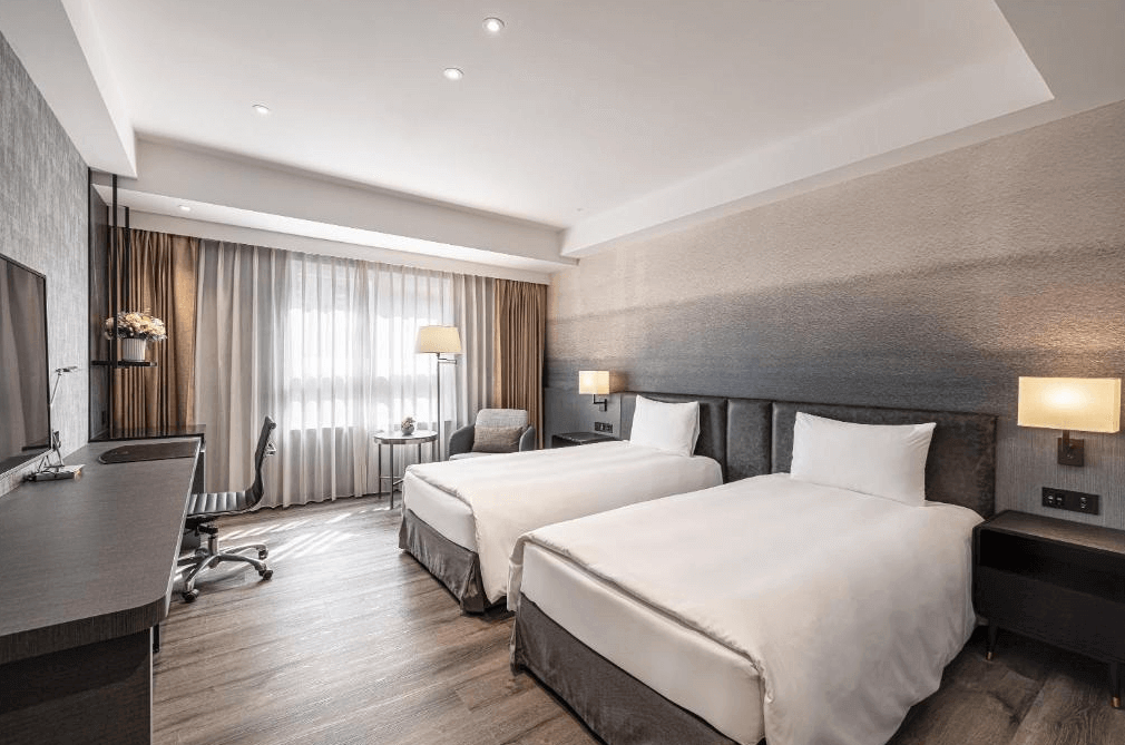 【新竹火車站住宿】煙波大飯店 新竹都會館的房間內設有兩張單人床，床頭牆面採用漸層灰色設計，搭配柔和的燈光，營造溫暖舒適的氛圍。房間配備木質地板、書桌與辦公椅，並設有大片落地窗，提供良好的採光。