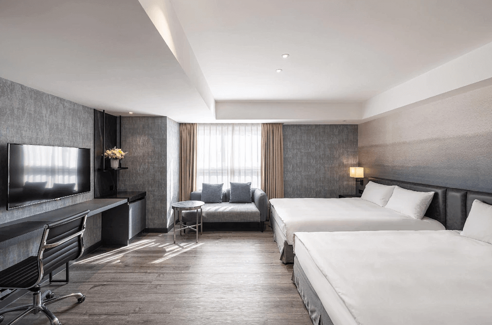 【新竹火車站住宿】煙波大飯店 新竹都會館寬敞的家庭客房內設有兩張雙人床，床頭牆面為灰色漸層設計，搭配現代感十足的家具。房間內設有一張灰色沙發和小型圓桌，提供休息與用餐的空間。大片窗戶讓自然光線充滿整個房間。