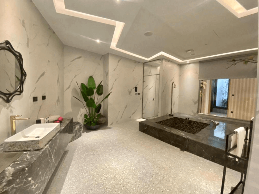 【新竹火車站住宿】琺何精品旅館的浴室空間寬敞，牆面採用大理石紋設計，中央設有一個黑色大理石浴缸，帶有簡約的現代感。洗手台與浴缸區域分開，搭配綠植裝飾，讓空間更具生活感與自然氛圍。浴室內的柔和燈光增添溫暖氣息。