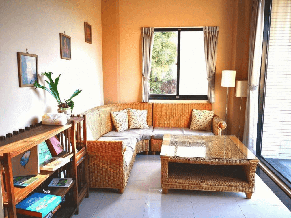 【埔里住宿】白舍·青丘的客廳角落，設有藤編沙發與透明玻璃茶几，牆面掛有多幅裝飾畫，旁邊擺放著書架和綠植，窗戶提供柔和的自然光，營造出舒適的居家氛圍。