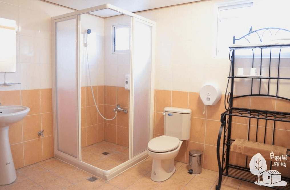 【嘉義寵物友善住宿】竹崎白樹腳驛棧的浴室，配有淋浴間、馬桶和洗手台，牆面與地板以米色瓷磚鋪設，旁邊放置一個黑色鐵製收納架，設計簡單實用。
