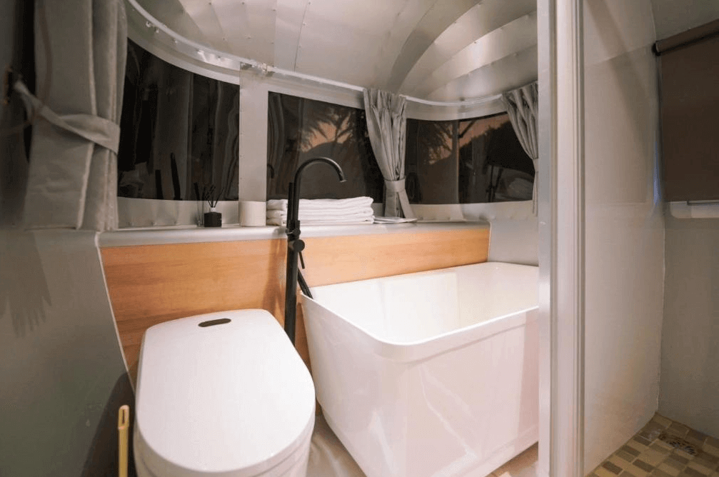 【埔里住宿】緣溪行森林營地的露營車內的浴室，設有白色浴缸、馬桶與黑色水龍頭，窗戶提供自然光線，木質與金屬材質的結合呈現簡約而時尚的設計風格。