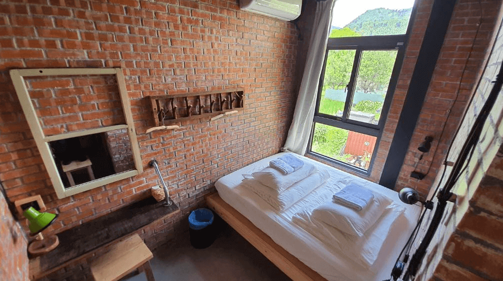 【埔里住宿】缽割貓點點的磚牆為主題的雙人房，設有一張木質基座的床鋪，床上整齊擺放著白色床單和毛巾。房間內部還有木製桌椅和簡約的擺設，窗外景色為綠意盎然的自然風光，整體設計給人溫暖復古的感覺。
