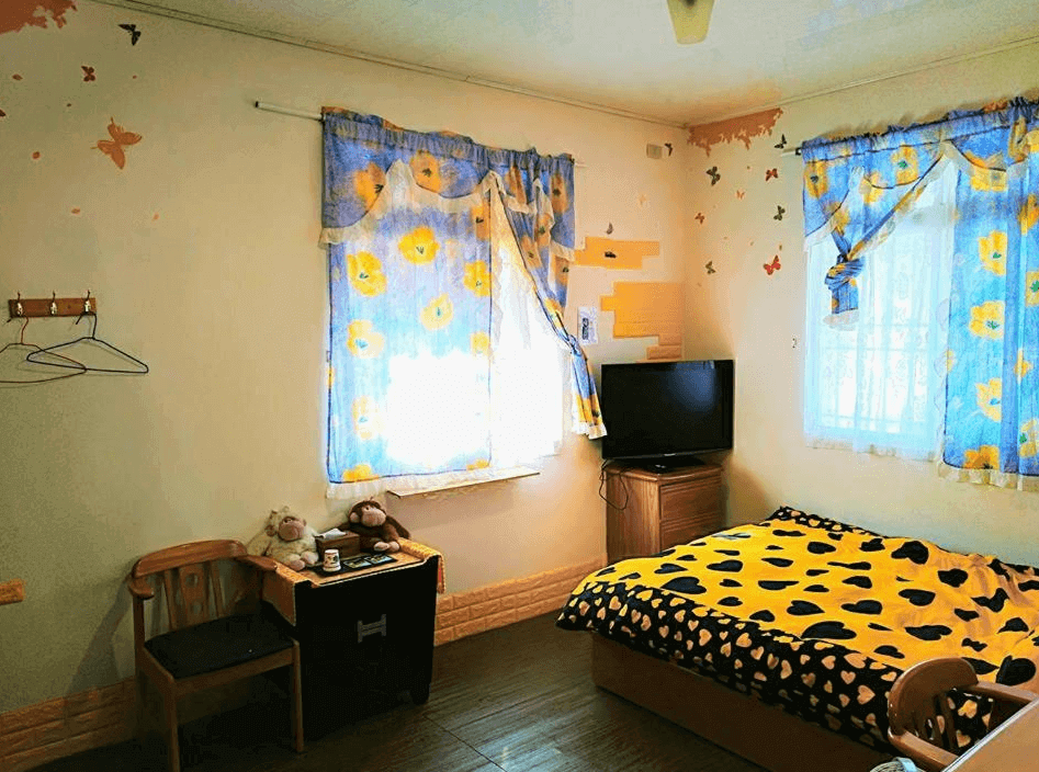 【廬山溫泉】翔婷茶工坊-登坊輕生活的風格樸實的單人房，以黃色與藍色花卉窗簾裝飾，牆面帶有蝴蝶與手繪圖案，呈現活潑溫馨的氛圍。房內配備電視、小木桌與布偶擺飾，床上鋪有豹紋與愛心圖案的暖色系被單，展現溫暖的住宿風格。