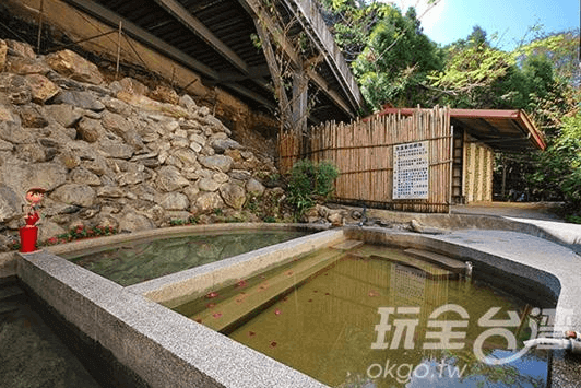 【廬山溫泉】翔婷茶工坊-登坊輕生活的戶外石砌溫泉池，環境自然靜謐，周圍有竹籬笆與石牆，展現純樸的鄉村風情。池水清澈，漂浮著紅色花瓣，營造出放鬆療癒的泡湯體驗。遠處可見溫泉區的簡約更衣室建築。