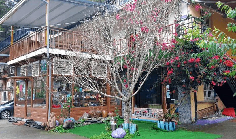 【廬山溫泉】翔婷茶工坊-登坊輕生活的兩層樓的木質建築，周圍綠意盎然，庭院內種有紅花樹與各種植栽。建築一樓設有透明玻璃窗，可見內部擺設手作藝品與茶具，呈現溫馨的茶坊風格。戶外鋪設綠色人工草皮，擺放可愛擺飾，增添田園氛圍。