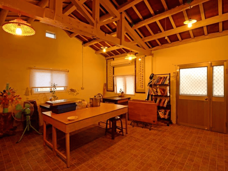 【嘉義寵物友善住宿】船仔頭天賞居民宿古樸風格的公共用餐區，內部配有木製餐桌和書架，天花板為裸露的木樑設計，牆上掛有書法卷軸，燈光柔和，環境寧靜。