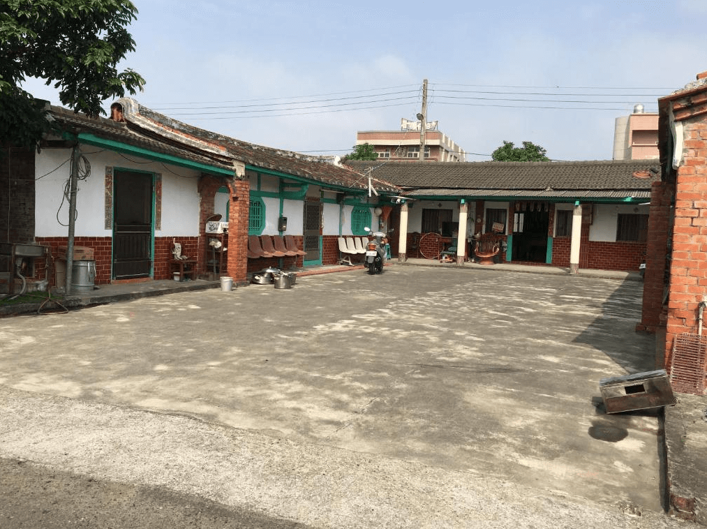 【嘉義寵物友善住宿】船仔頭天賞居民宿的台灣三合院建築，紅磚牆搭配綠色木框門窗，中庭空間寬敞，地面為水泥鋪設，院內擺放著一些農具和舊式家具，充滿濃厚的鄉村氣息。