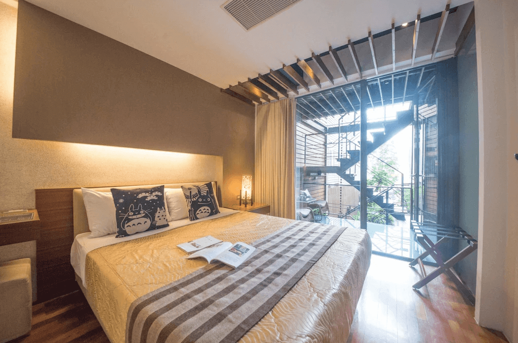 【日月潭住宿】苔米屋的房間以溫暖的木質地板與牆面搭配，設計融合日式簡約與現代感。床頭燈採用創意木格設計，增添質感與立體感。玻璃門外可見旋轉樓梯與綠意盎然的戶外景觀，充滿自然氣息。