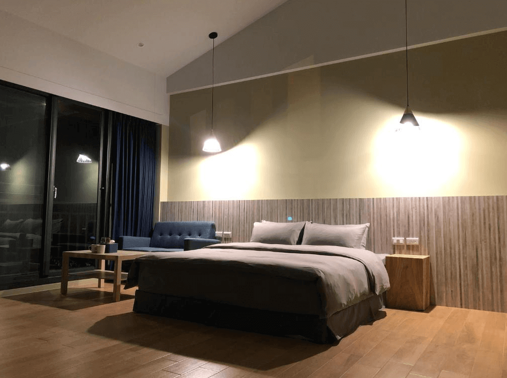 【阿里山住宿】茶緣觀舍昏黃的燈光營造出溫暖舒適的環境，房內擺設包括一張大床與休息沙發，適合享受寧靜的夜晚。牆面的木紋設計與吊燈相得益彰，展現溫馨的住宿氛圍。