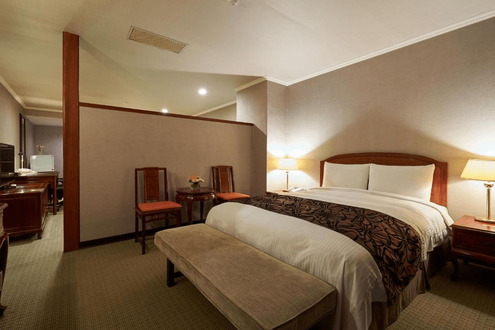 【新竹火車站住宿】迎曦大飯店的房間內設有一張雙人床，搭配木質床頭與家具，呈現古典優雅的風格。床尾設有一張長凳，兩側擺放檯燈，營造溫馨的光線。房間內還配有一張小圓桌與兩張椅子，提供旅客舒適的休息空間。