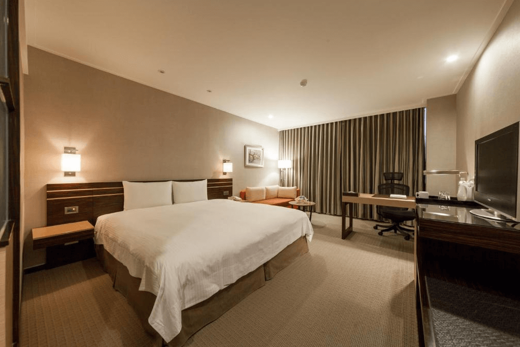 【新竹火車站住宿】迎曦大飯店的房間內設有一張大雙人床，搭配白色床品與木質家具，風格簡約時尚。房間內配置了辦公桌、椅子以及沙發區，適合商務旅客使用。大片窗簾搭配柔和燈光，讓空間顯得明亮舒適。