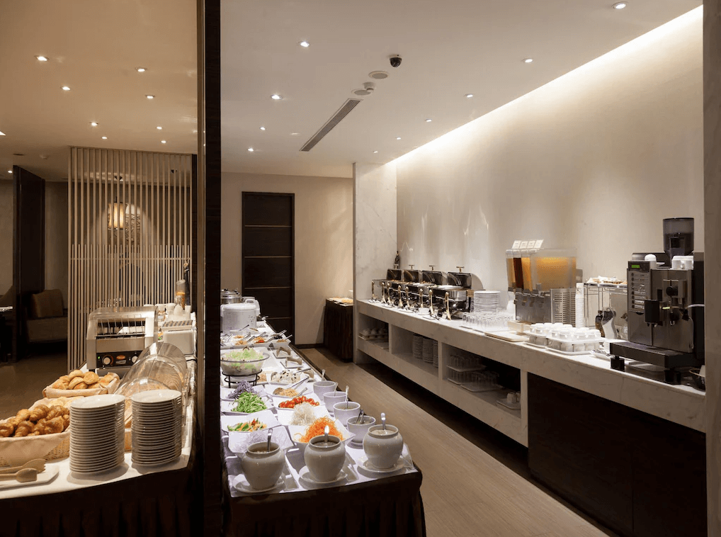 【新竹火車站住宿】迎曦大飯店的自助餐廳內設有多種類型的餐食，包括麵包、水果、沙拉與熱食，陳列在整齊的餐台上。空間採用溫暖的燈光與木質裝飾，營造出溫馨的用餐氛圍，適合旅客開始一天的美好時光。