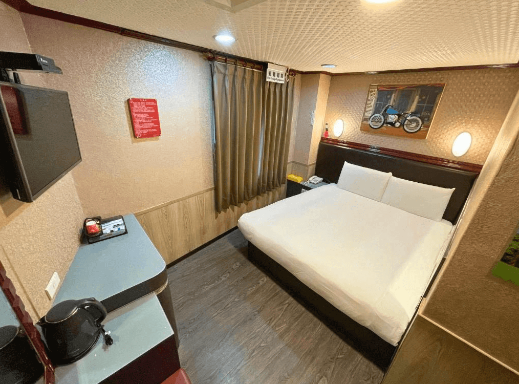 【寧夏夜市住宿】迪樂商旅的雙人房，設有大床，牆面為暖色調裝飾，配有懸掛電視、電熱水壺與小型工作台，窗戶設有灰色窗簾，營造簡潔實用的空間。