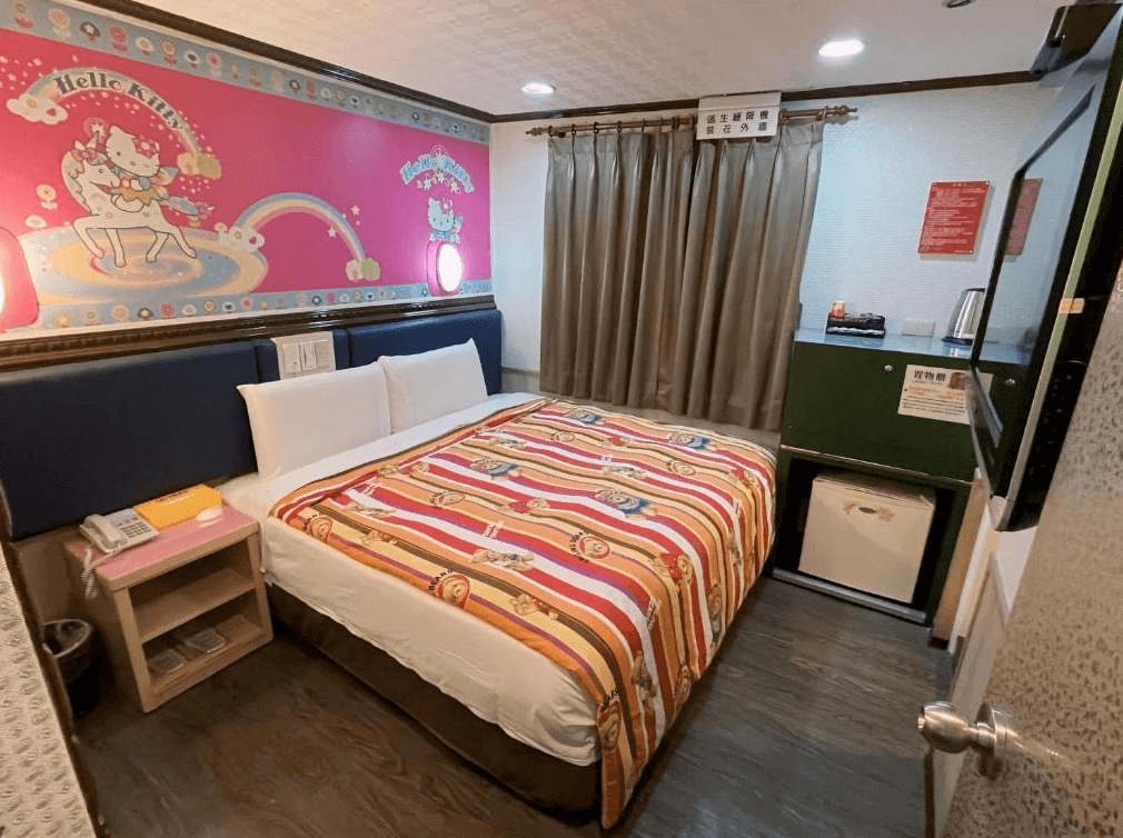 【寧夏夜市住宿】迪樂商旅的雙人房，牆面有粉紅色的 Hello Kitty 圖案壁畫，床鋪上鋪有卡通圖案的床單，房內設有小冰箱、電視及基本生活設施。