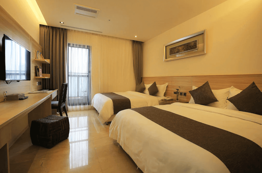 【新竹火車站住宿】達龍商旅的房間內設有兩張雙人床，床鋪整潔舒適，配有棕色抱枕與被套裝飾。窗戶採用大面積落地設計，透入自然光線，搭配淺色窗簾，整體氛圍明亮溫馨。左側設有書桌與壁掛式電視，並配有幾本書，營造出實用且舒適的住宿環境。