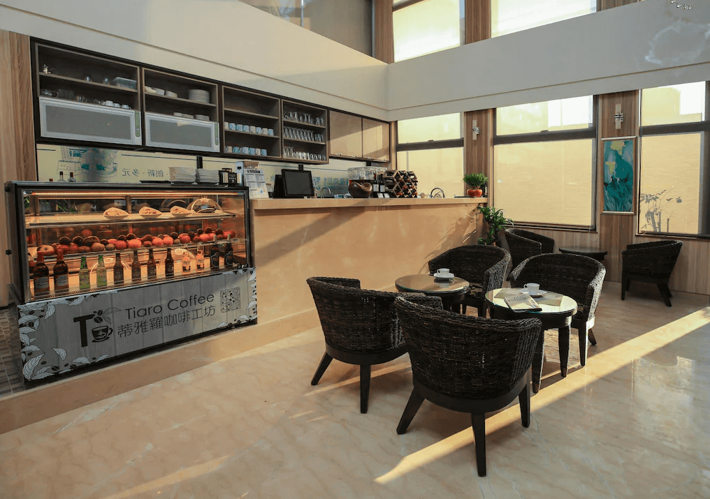 【新竹火車站住宿】達龍商旅的咖啡廳，設有多張藤編椅與小圓桌，適合旅客休憩或品嚐咖啡。櫃檯上方陳列著多種飲品和甜點，背景牆的開放式展示架上擺放著杯具，空間採用淺木色調與充足的自然光線，營造出舒適的用餐氛圍。